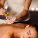 Frau auf dem Bauch liegend, bereit für eine Massage. Hinter ihr eine Frau, die sich gerade Öl für die Massage in ihre Hand gießt.